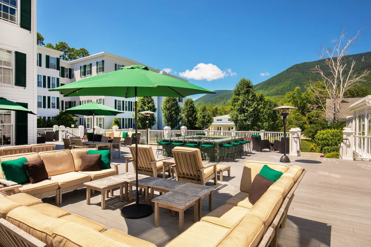 An inviting outdoor seating area under green umbrellas offers panoramic mountain views. Guests can be seen relaxing and socializing, evoking a leisurely atmosphere ideal for morning breakfasts, sunset cocktails, or al fresco dining. The scene radiates the relaxed luxury and natural beauty that defines New England escapes.