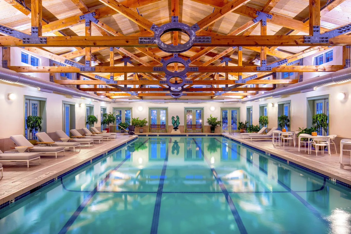 A serene indoor swimming pool space featuring elegant wooden beams, comfortable lounge chairs, a striking sculpture at the far end, and expansive windows letting in soft natural light. The pool is inviting, with a tranquil ambiance ideal for morning laps or relaxing swims. The setup evokes a refined resort atmosphere, and the area is spacious enough for yoga sessions or small social gatherings.