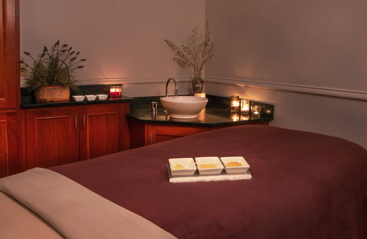 A serene spa setting with a massage table, decorative bowls, candles, and a sink, evoking relaxation and wellness. The soft lighting and soothing decor reinforce a sense of indulgence and tranquility, reminiscent of luxurious spa treatments.