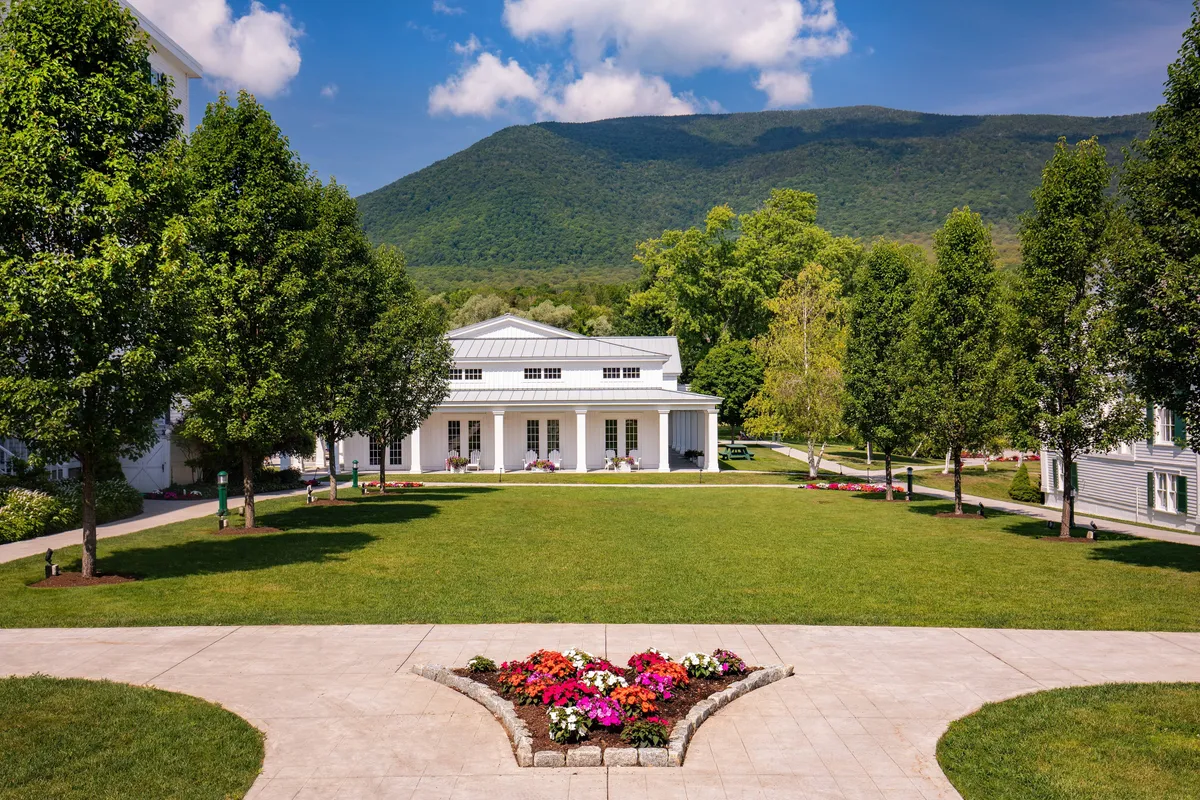 This exterior image features the white main resort building adorned with stately columns, set against a manicured lawn, vibrant flower beds, lush trees, and the dramatic Green Mountains in the background. The setting is peaceful and inviting, with visible walking paths perfect for evening strolls and multiple outdoor gathering spots ideal for social events or relaxation.