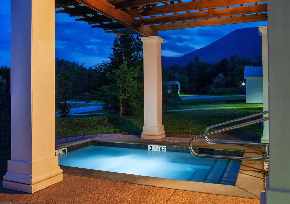 An inviting outdoor pool area illuminated under the evening sky, with a pergola structure and a breathtaking mountain backdrop. The pool glows softly, suggesting a soothing environment for evening swims or stargazing. The setting emphasizes relaxation, luxury, and connection with the outdoors, enhancing the sense of an all-encompassing resort experience.