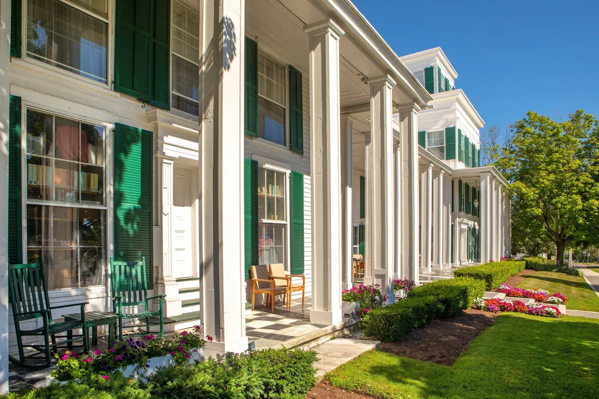 This exterior image features a charming porch with green shutters, classic rocking chairs, blooming flower beds, and stately columns. The presence of people relaxing on the porch and tending to the garden creates a lively, communal atmosphere. The scene is warmly lit, suggesting a vibrant, welcoming setting ideal for socializing after outdoor adventures.