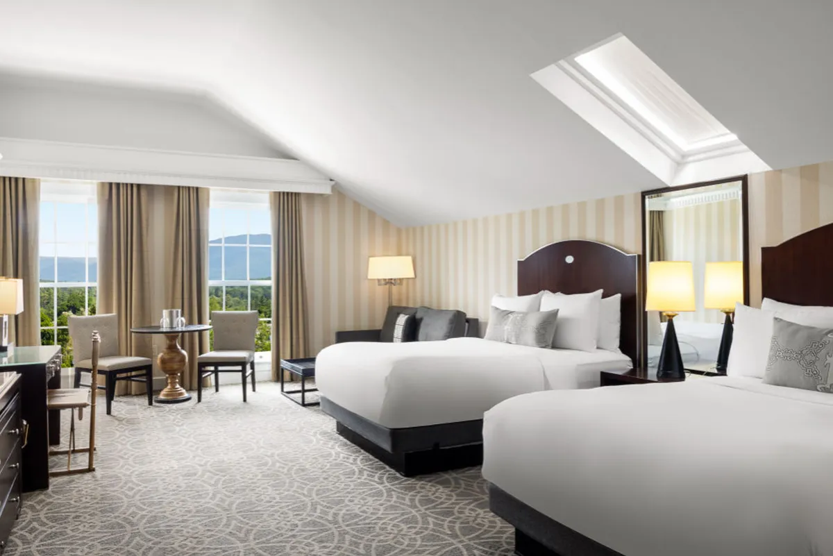 The image features a serene studio interior with two plush beds, a large window revealing peaceful mountain views, a cozy sitting area with a table and chairs ideal for morning coffee or planning the day, and warm, decorative lighting that enhances the tranquil atmosphere.